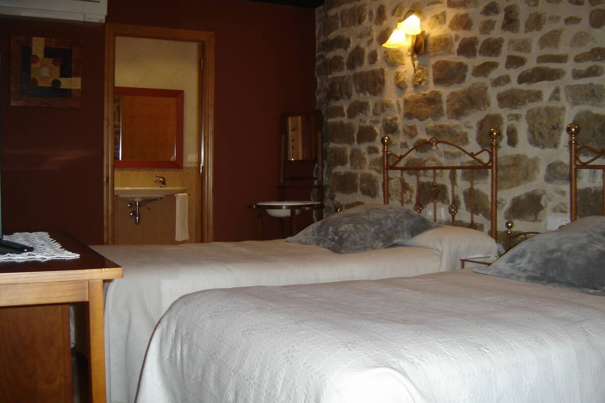 Ein Zimmer mit zwei Betten, einem Holztisch, einer Steinwand und einem durch eine offene Tür einsehbaren Badezimmer.