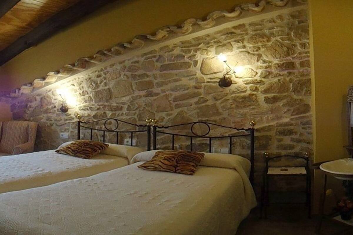 Ein Schlafzimmer mit zwei Betten, einer Steinwand, einer Holzbalkendecke, einem Sessel und einem kleinen Tisch.