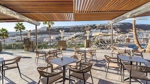 An outdoor terrace with wicker furniture, a pool, and a view of a coastal cityscape. - Cala Nova Apartamentos (Mogan)