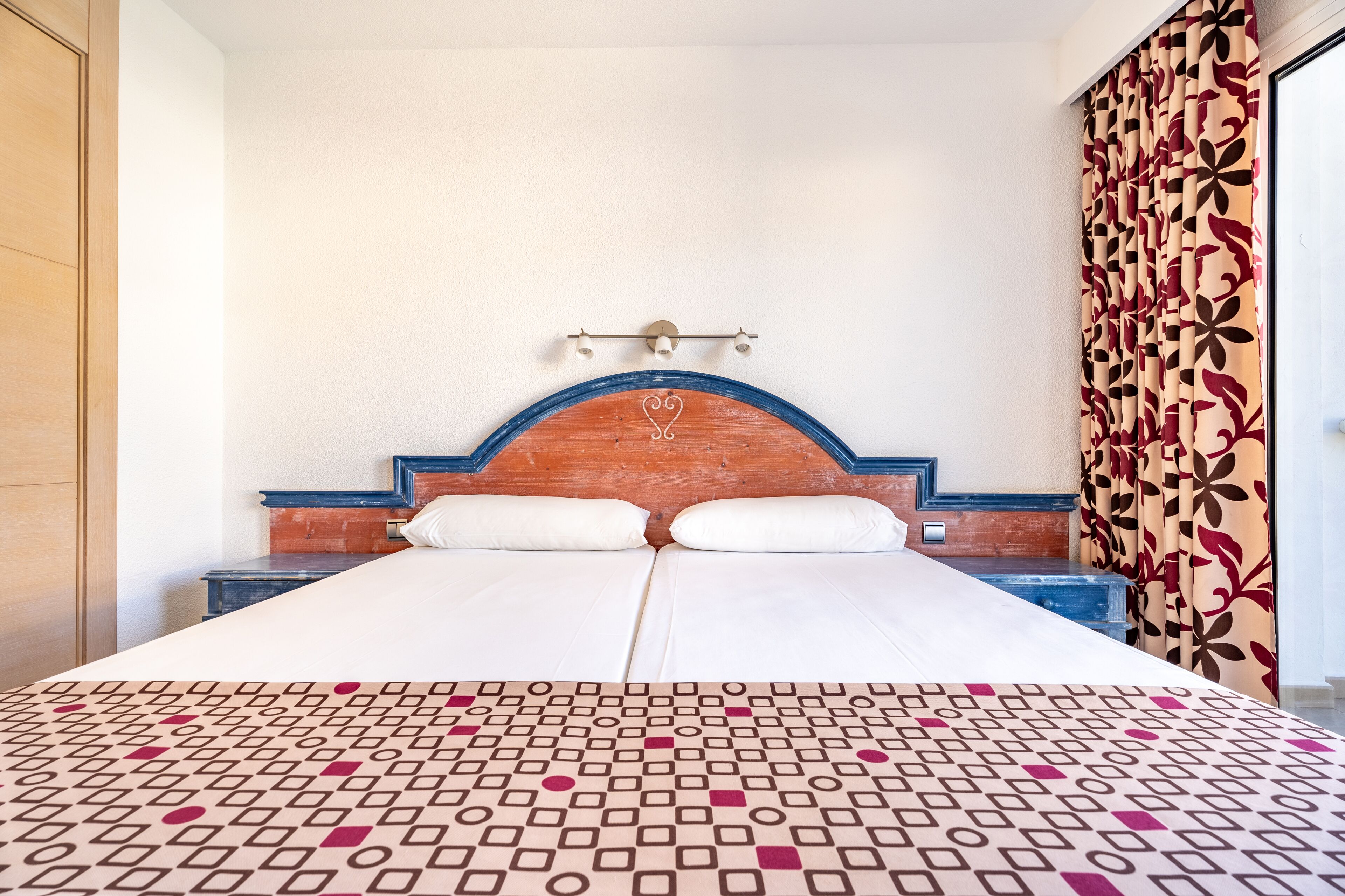 A hotel room with a large bed, a wooden headboard, a patterned bedspread, and a curtain with a floral design.