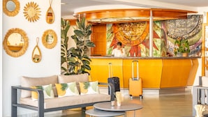 A hotel lobby with a sofa, two tables, a reception desk, and a colorful mural. - Cala Nova Apartamentos (Mogan)