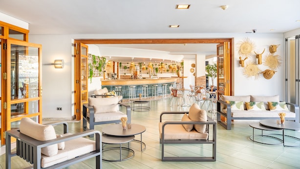 A spacious lounge area with modern furniture, a bar in the background, and a view of the city through large windows. - Cala Nova Apartamentos (Mogan)