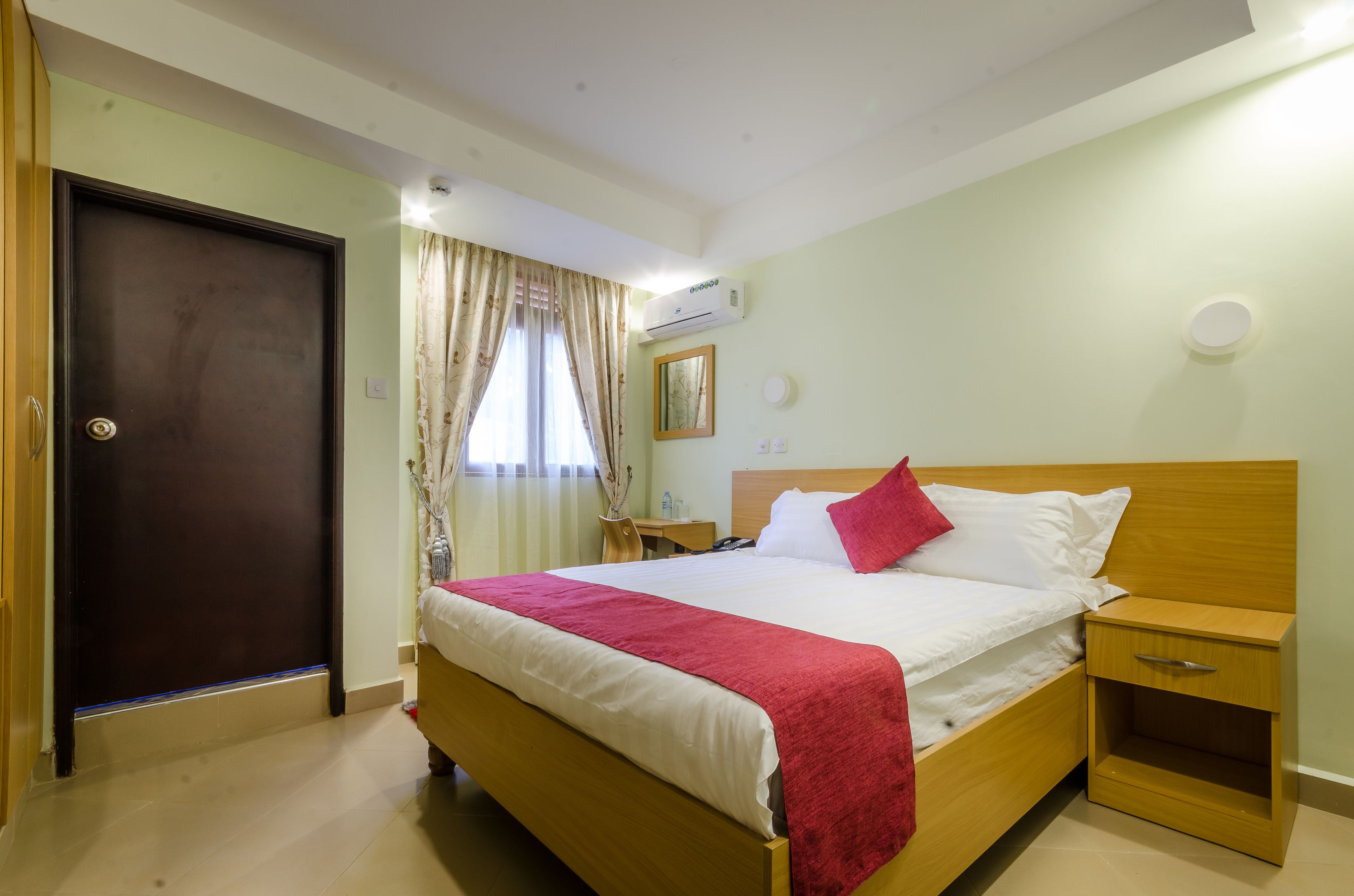 A hotel room with a bed, bedside table, nightstand, and an air conditioning unit.