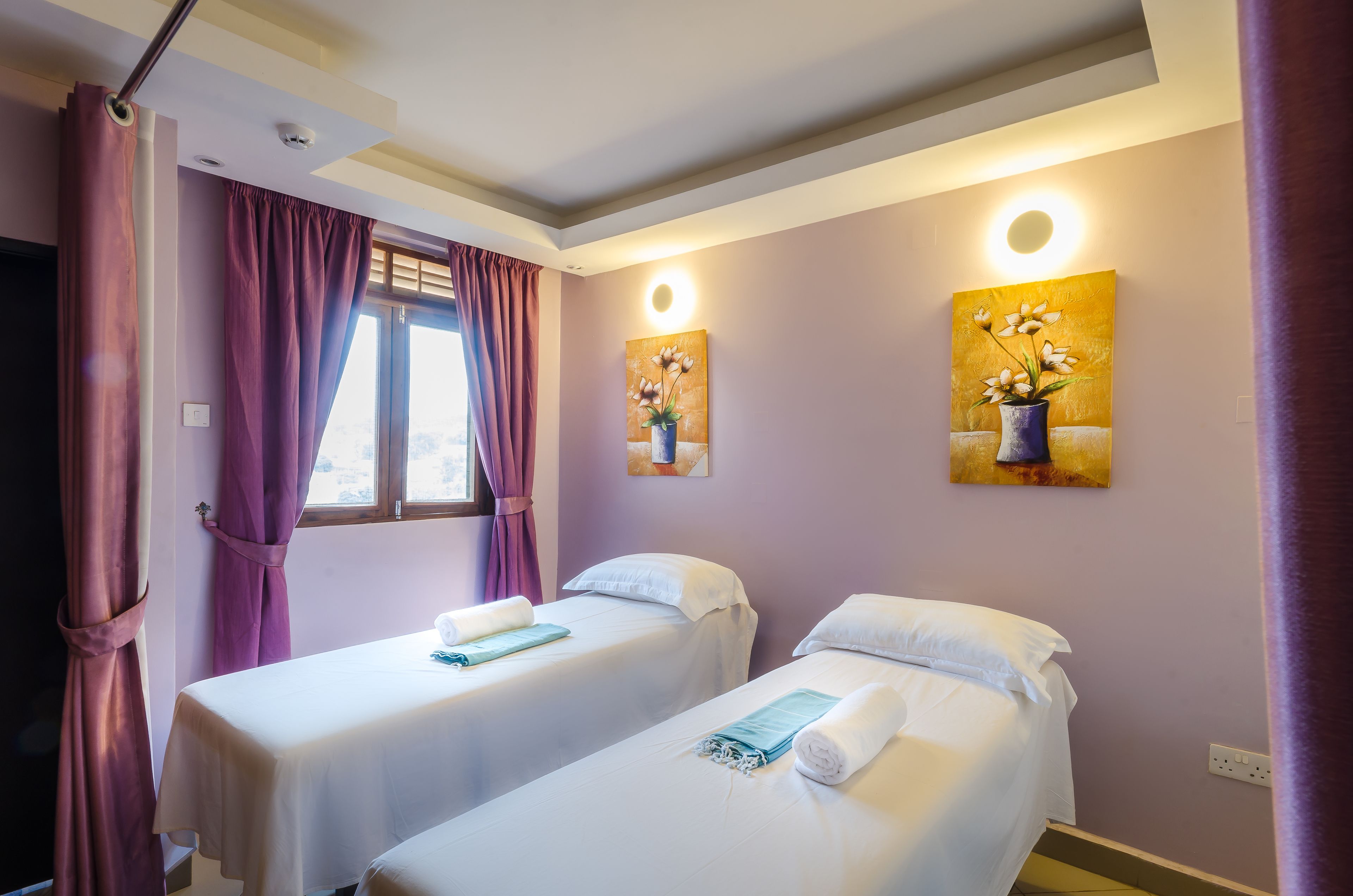 Une chambre d’hôtel avec deux lits, des rideaux violets et deux tableaux accrochés au mur.