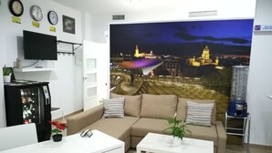 A living room with a sofa, a coffee table, a TV, a refrigerator, and a large cityscape mural on the wall.