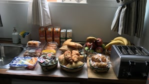 A breakfast setup with bread, pastries, fruits, and beverages on a table near a window.