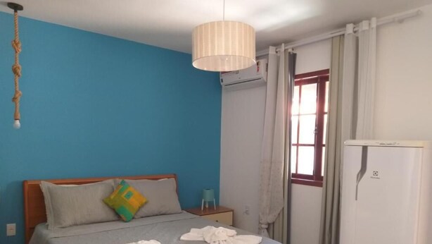 A bedroom with a bed, a bedside table, a hanging light fixture, a window with curtains, and a white refrigerator. - Pousada Melodia do Mar (Cabo Frio)