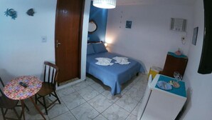 A small, compact room with a bed, a table, and a chair. There is a door, a wall-mounted mirror, and a ceiling light fixture. - Pousada Melodia do Mar (Cabo Frio)