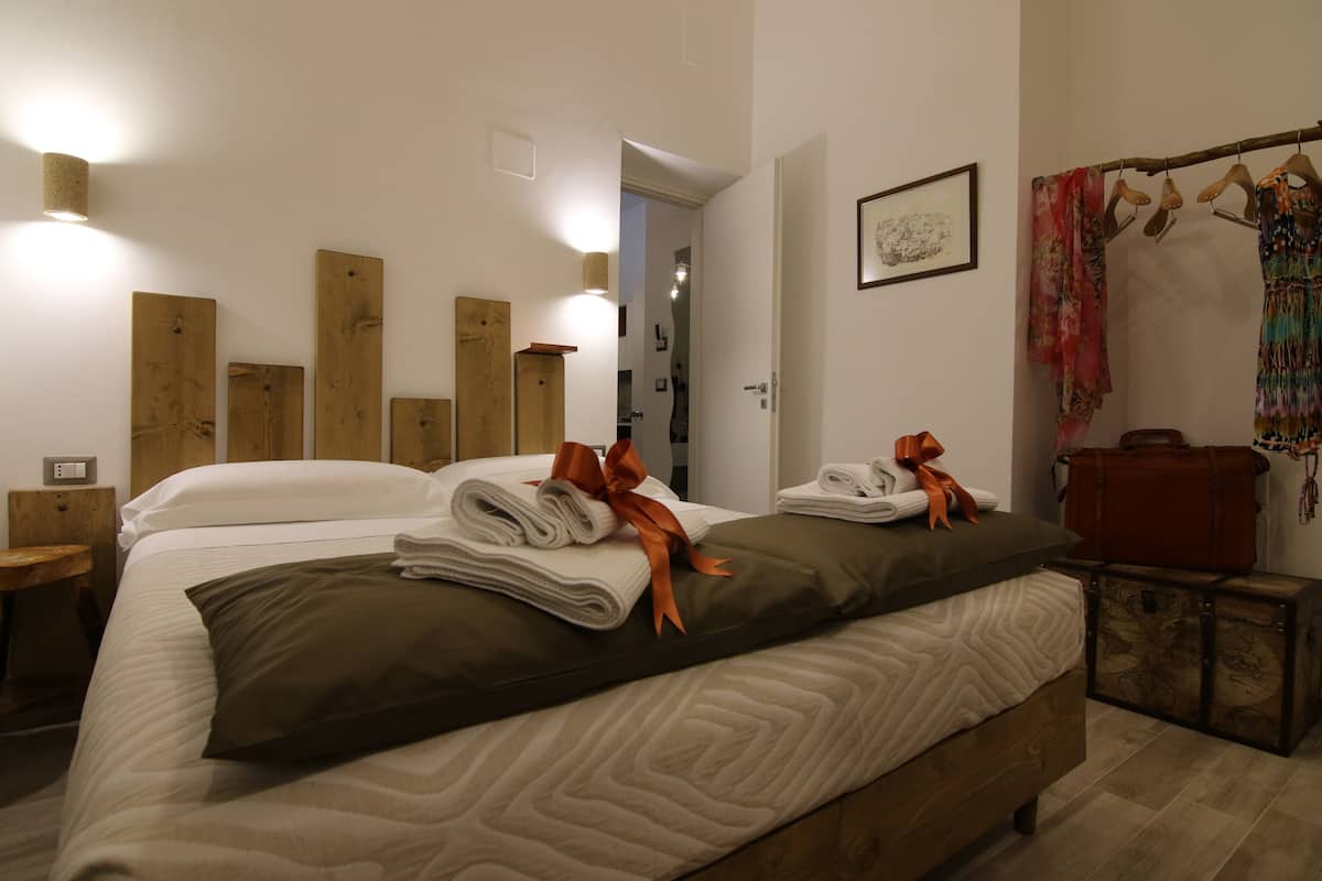 Een net opgemaakt bed met opgerolde handdoeken en lint, een houten hoofdbord, een klein houten nachtkastje, en een wandlamp.