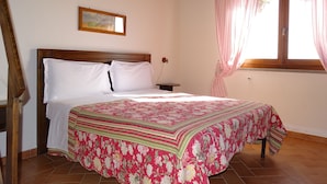A bedroom with a wooden bed frame, white bedding, a floral-patterned bedspread, a nightstand, a wall-mounted air conditioner, a window with pink curtains, and a painting above the bed. - Agriturismo Podere 1248 (Ladispoli)