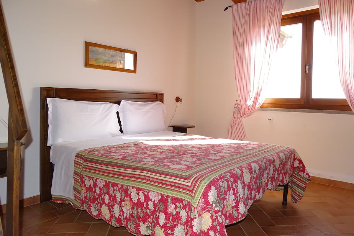 A bedroom with a wooden bed frame, white bedding, a floral-patterned bedspread, a nightstand, a wall-mounted air conditioner, a window with pink curtains, and a painting above the bed.