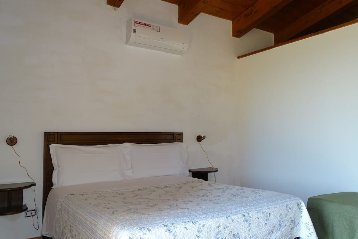 A bedroom with a wooden headboard, a bed with white bedding, a nightstand with a lamp, and a wall-mounted air conditioner.