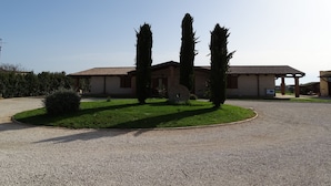 A building with a tiled roof, a gravel driveway, and two tall cypress trees in the foreground. - Agriturismo Podere 1248 (Ladispoli)