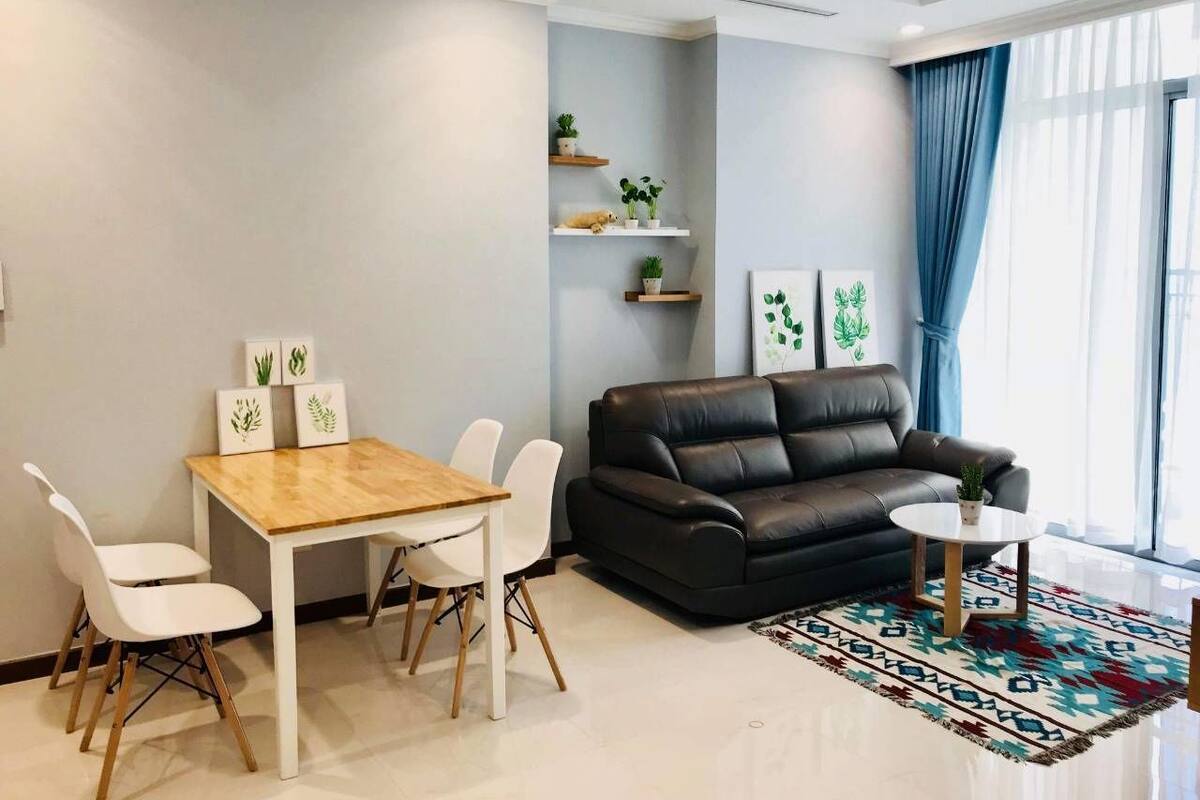 A modern living room with a black leather sofa, a wooden dining table with white chairs, and a colorful rug.