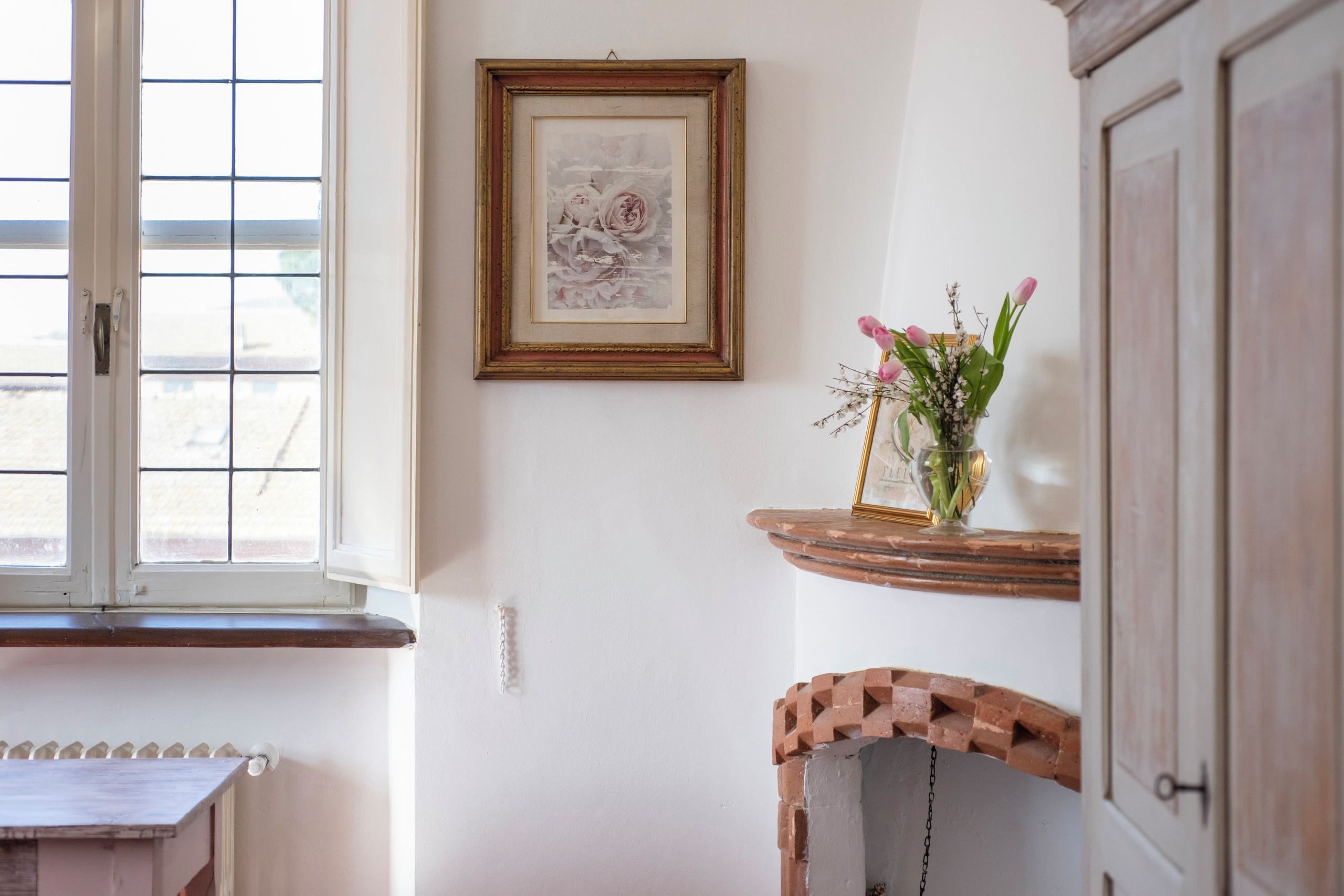 Une pièce avec une fenêtre, un tableau encadré au mur, un vase rempli de fleurs sur une étagère et une chaise dont l’assise est décorée d’un motif imitant des briques.