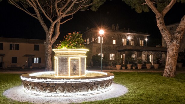 A stone fountain with a light-up base, surrounded by grass and trees, with a building in the background. - Fattoria Pian di Rocca (Castiglione della Pescaia)