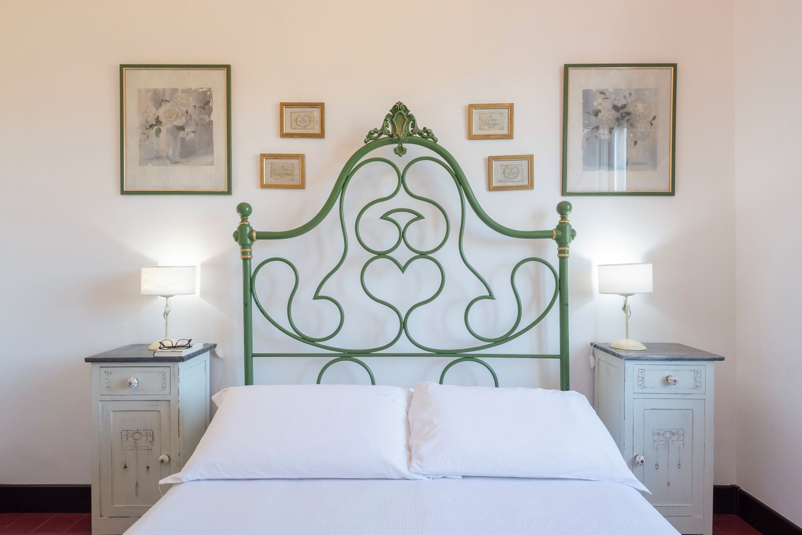A bed with a green metal headboard, two bedside tables with lamps, and framed pictures on the wall.