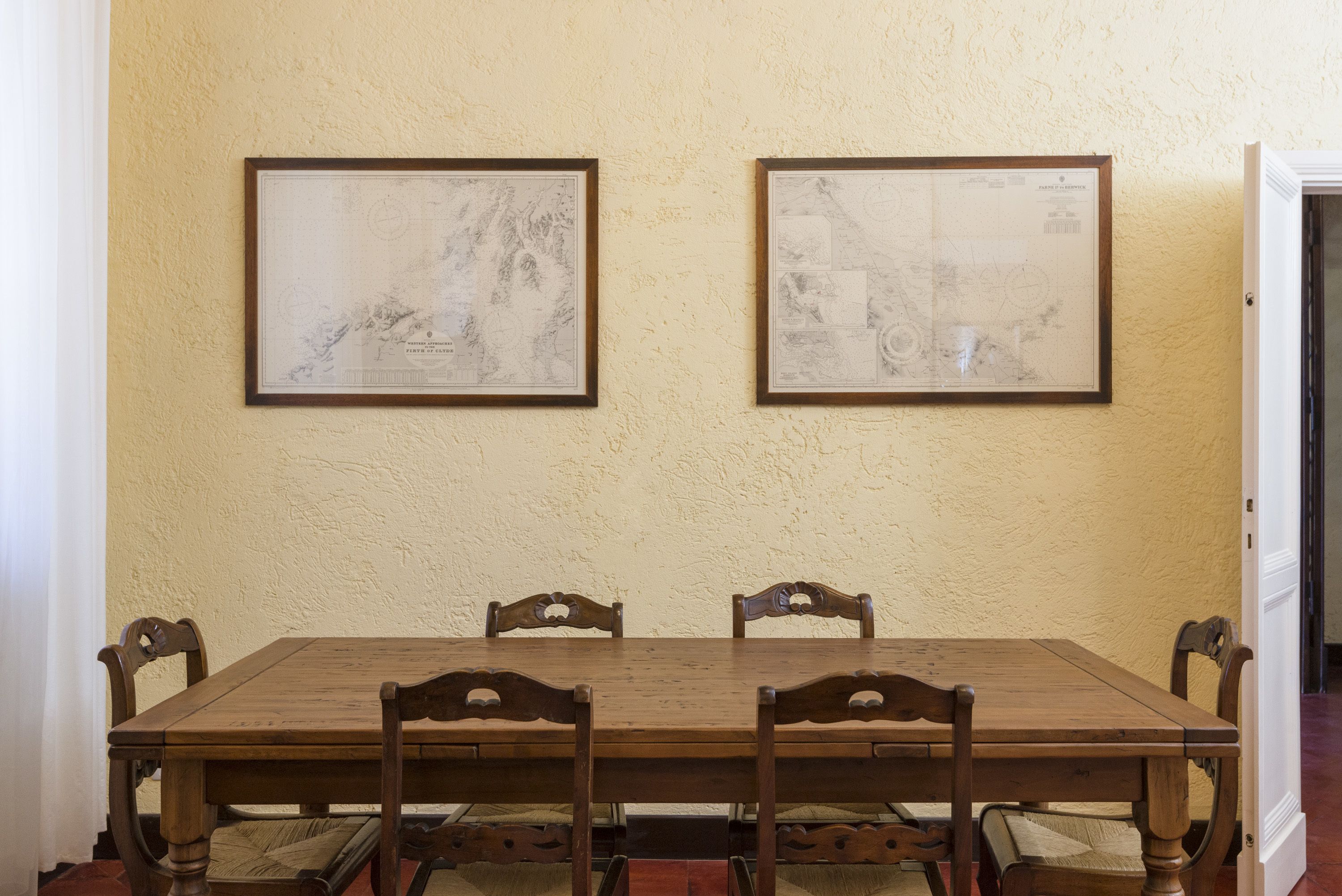A dining area with a wooden table and chairs, two framed maps on the wall, and a door to the right.