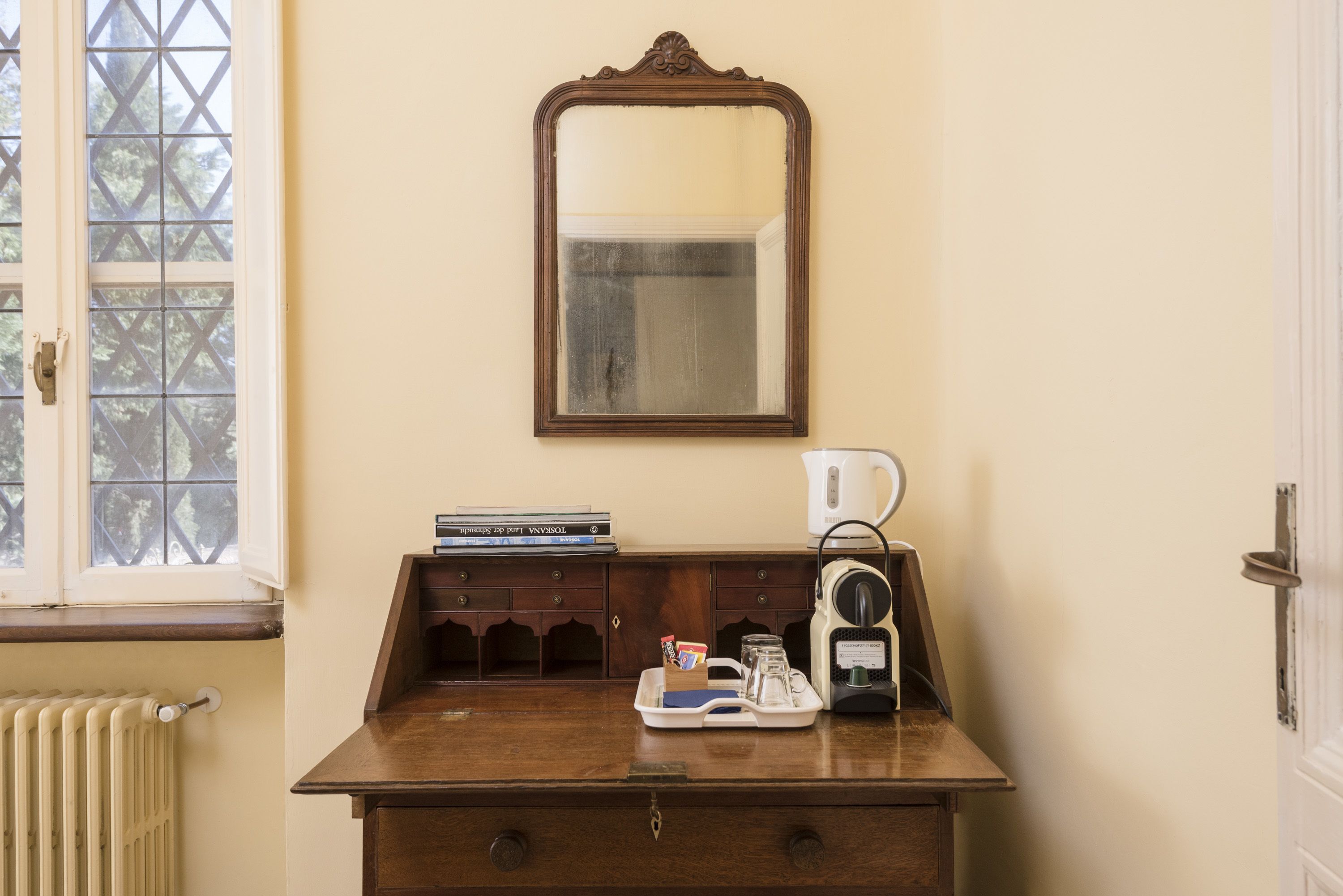 A room with a wooden dresser, a mirror, a radiator, a window with a view of trees, and a coffee maker on the dresser.