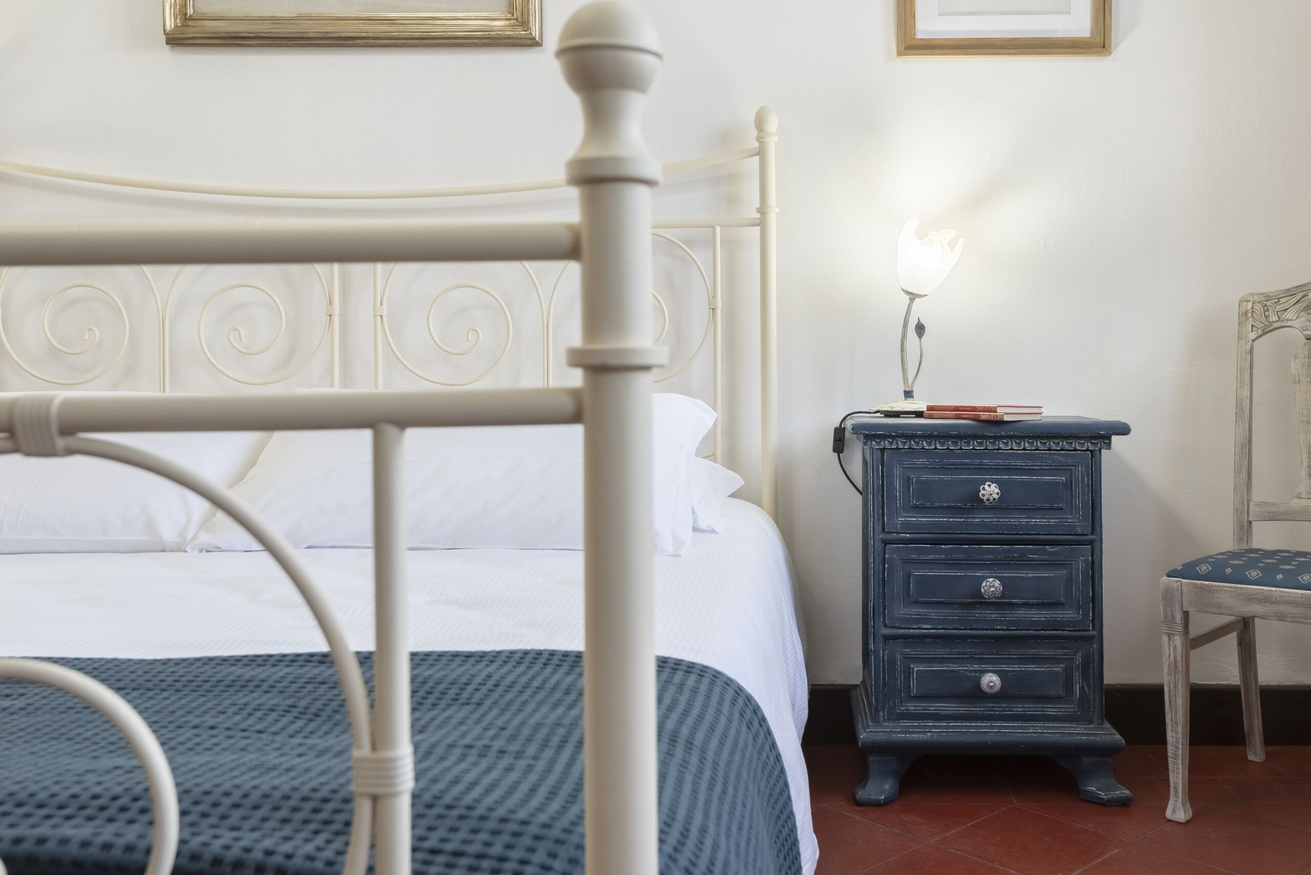 Una cama bien hecha con cabecera blanca, una mesita de noche azul con cajones y una silla con cojín estampado.
