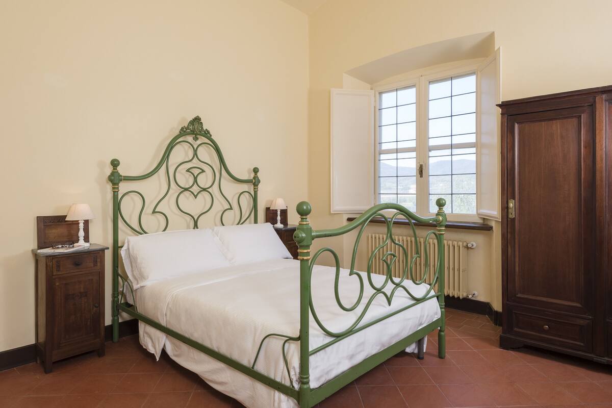 A single bed with a green metal frame, a wooden nightstand with a lamp, and a window with a view of the outside.