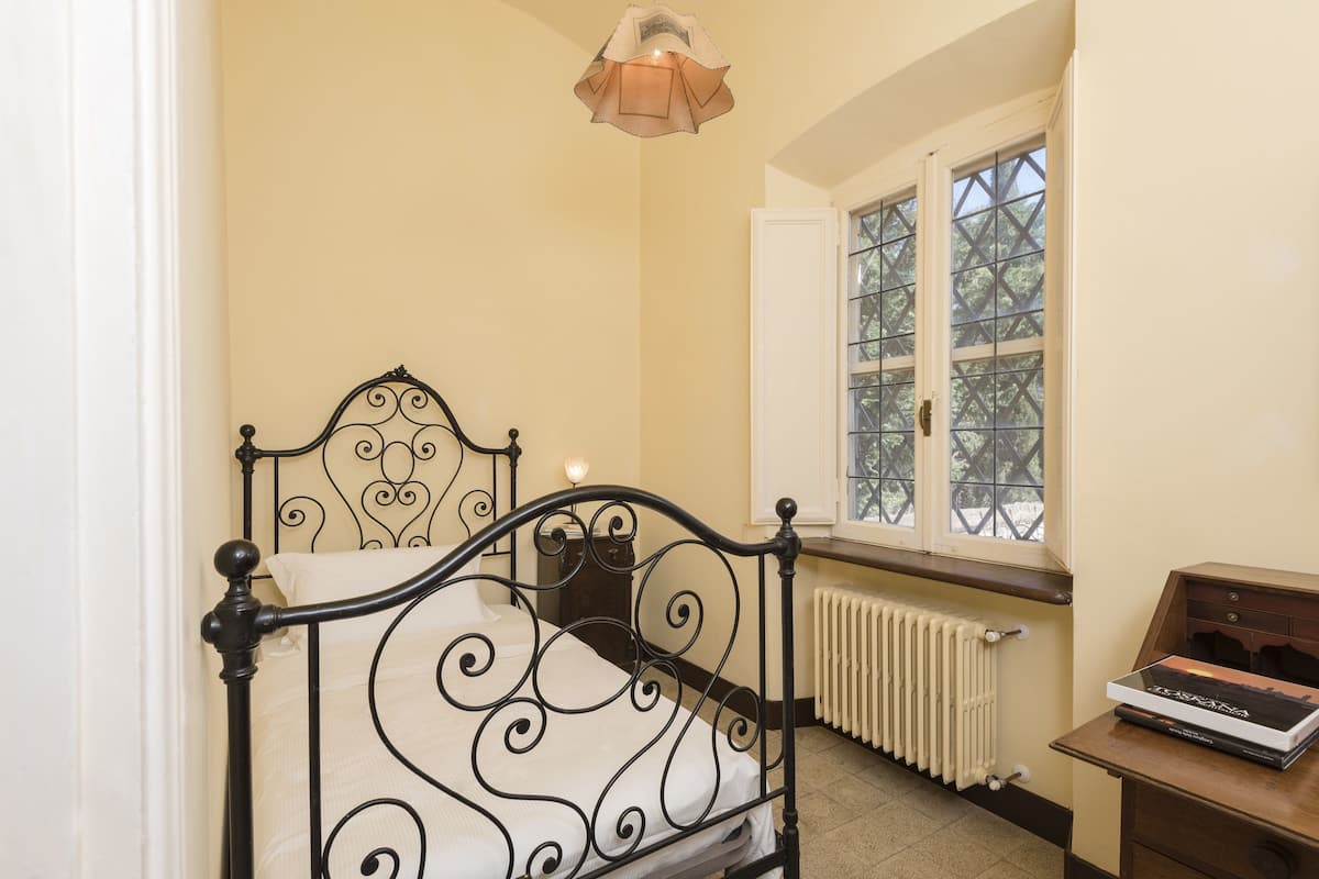 A bedroom with a bed, a radiator, a window with a view, and a desk with a book.