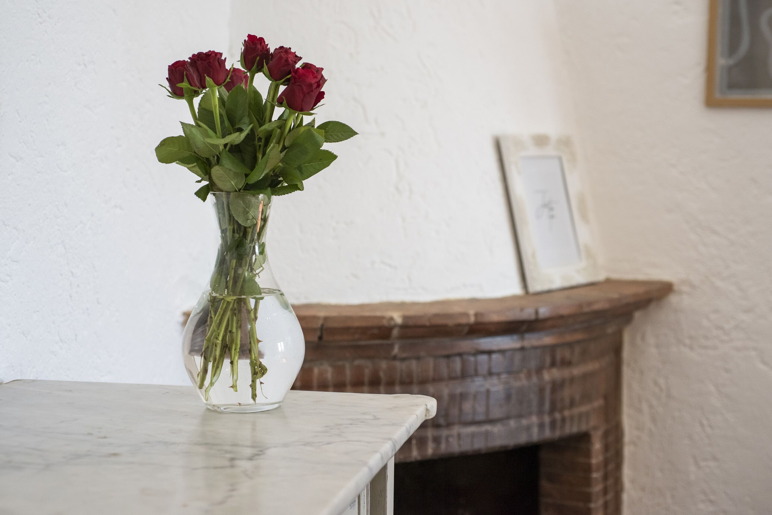 A clear vase with red roses on a marble table against a white wall with a framed picture and a brick fireplace.