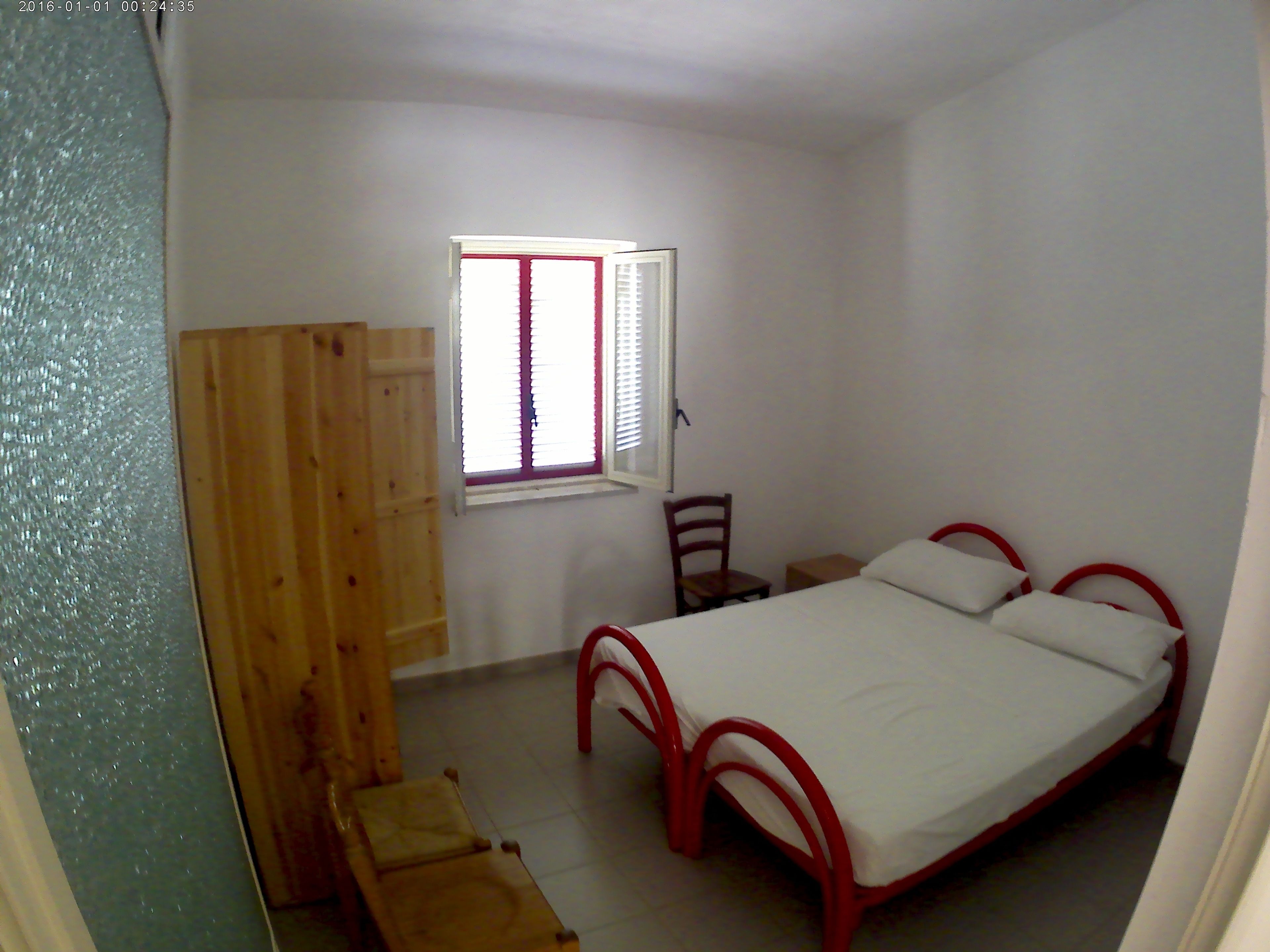 A small, simple bedroom with a single bed, a wooden chair, a small table, and a window with blinds.