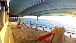 A beach with a playground, including a slide and climbing structures, under a shaded area.