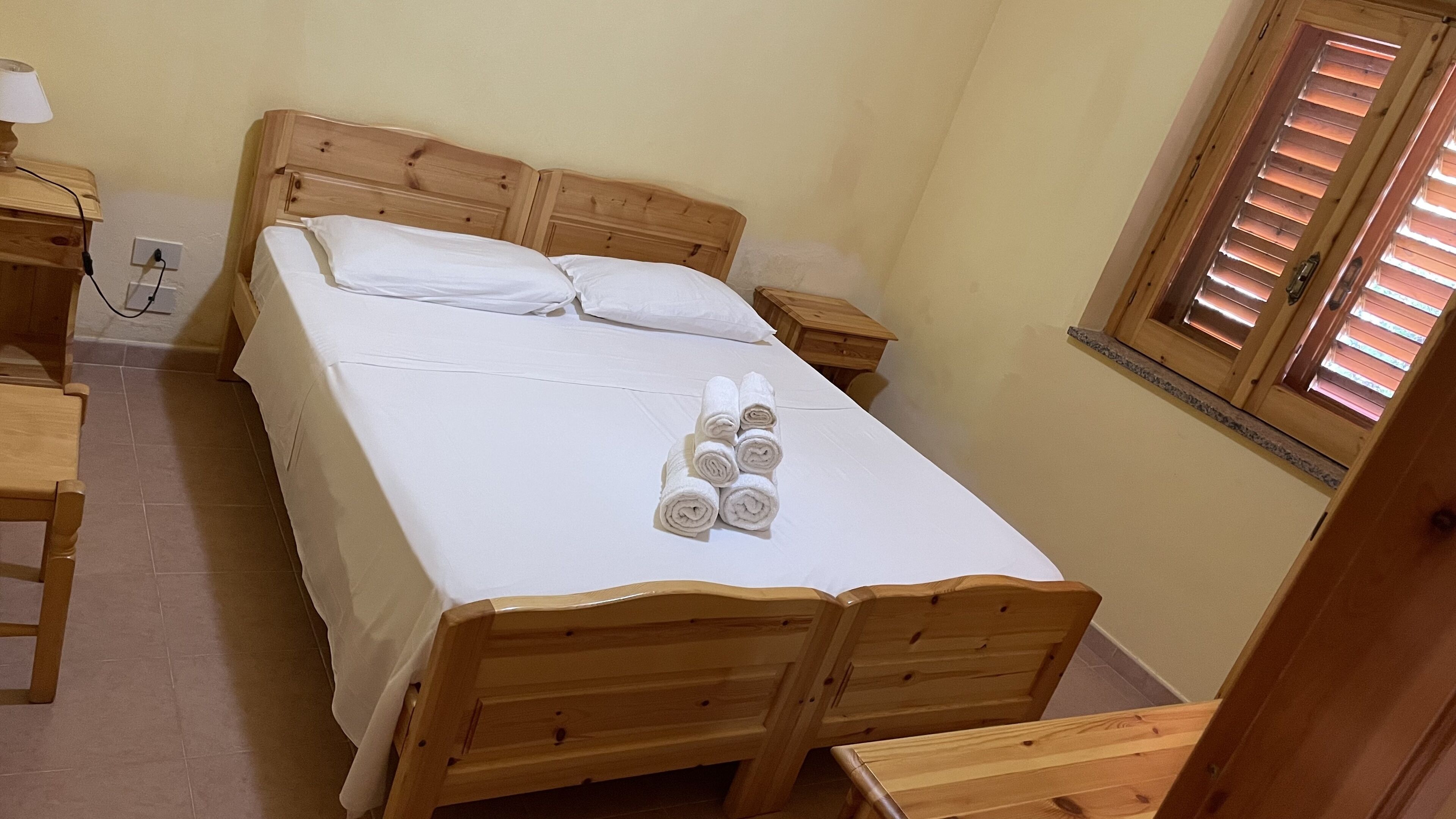 Una cama de madera con sábanas blancas, una mesita de noche y una ventana con persianas.