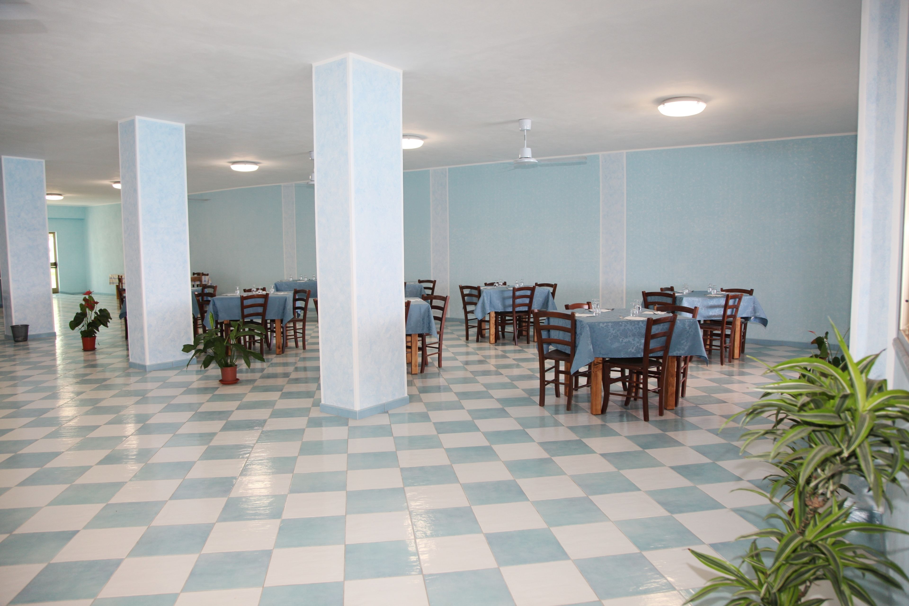 Un comedor amplio con suelo de cuadros, varias mesas con manteles azules, sillas de madera y plantas en macetas.