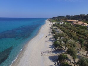 Una playa costera con aguas azules cristalinas, una orilla arenosa y una hilera de edificios con un paisaje montañoso al fondo.