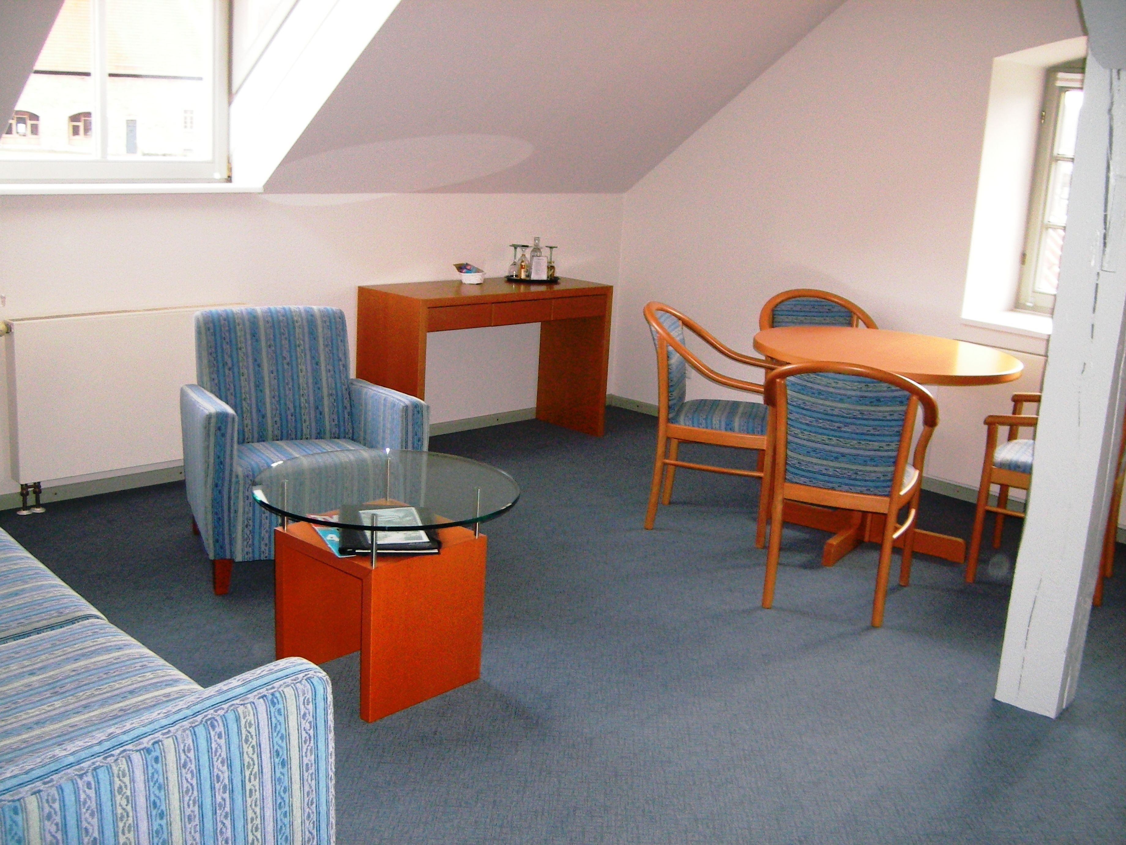 A small, cozy room with a sloped ceiling, a glass-top coffee table, a sofa, two striped armchairs, a wooden dining table with chairs, and a small side table.