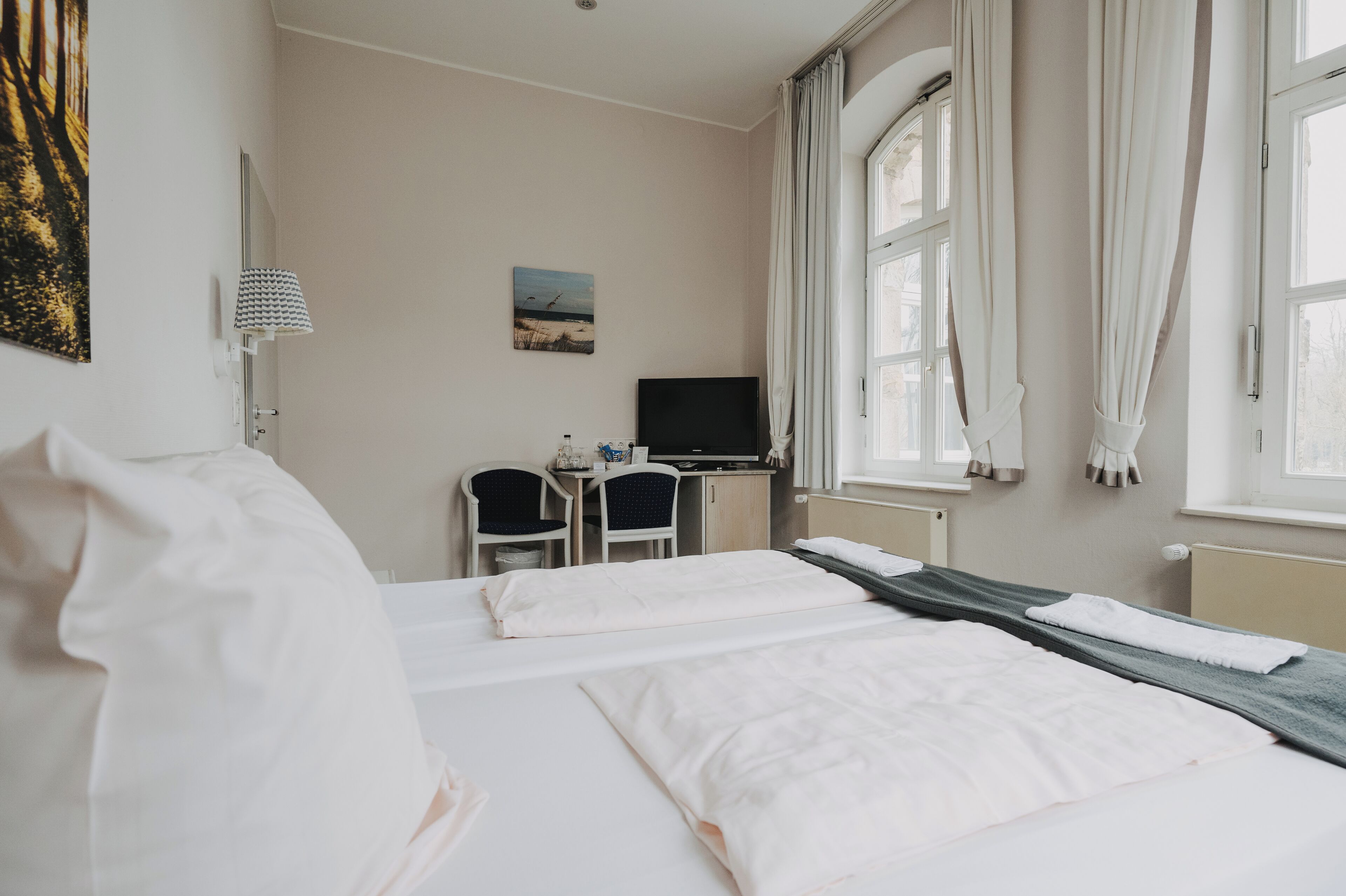 Ein Hotelzimmer mit zwei Betten, einem Schreibtisch, einem Stuhl, einem Fernseher und großen Fenstern mit Vorhängen.