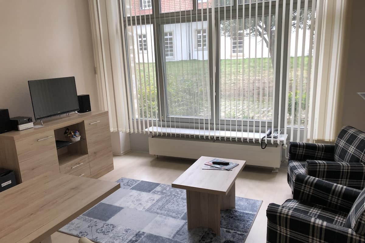 A modern living room with a sofa, a coffee table, a TV on a wooden cabinet, and a window with blinds.