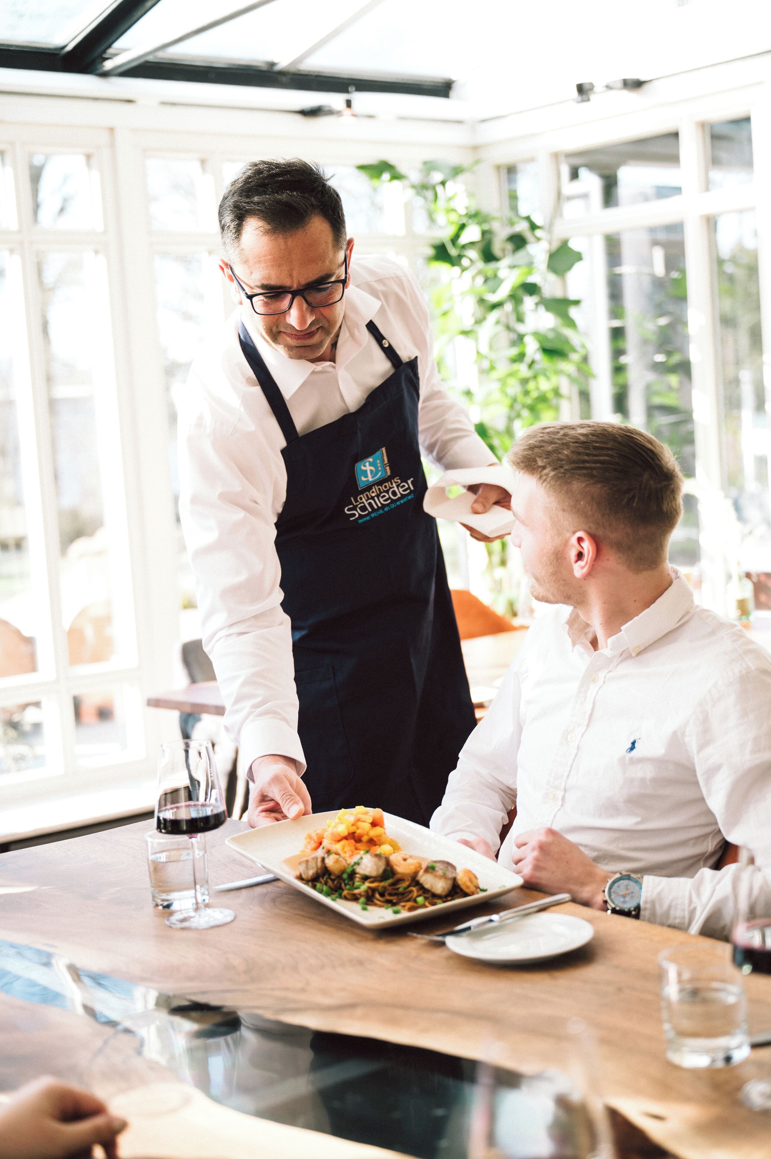 Ein Kellner serviert einem Gast eine Mahlzeit in einem hellen Restaurant mit großen Fenstern und Zimmerpflanzen.
