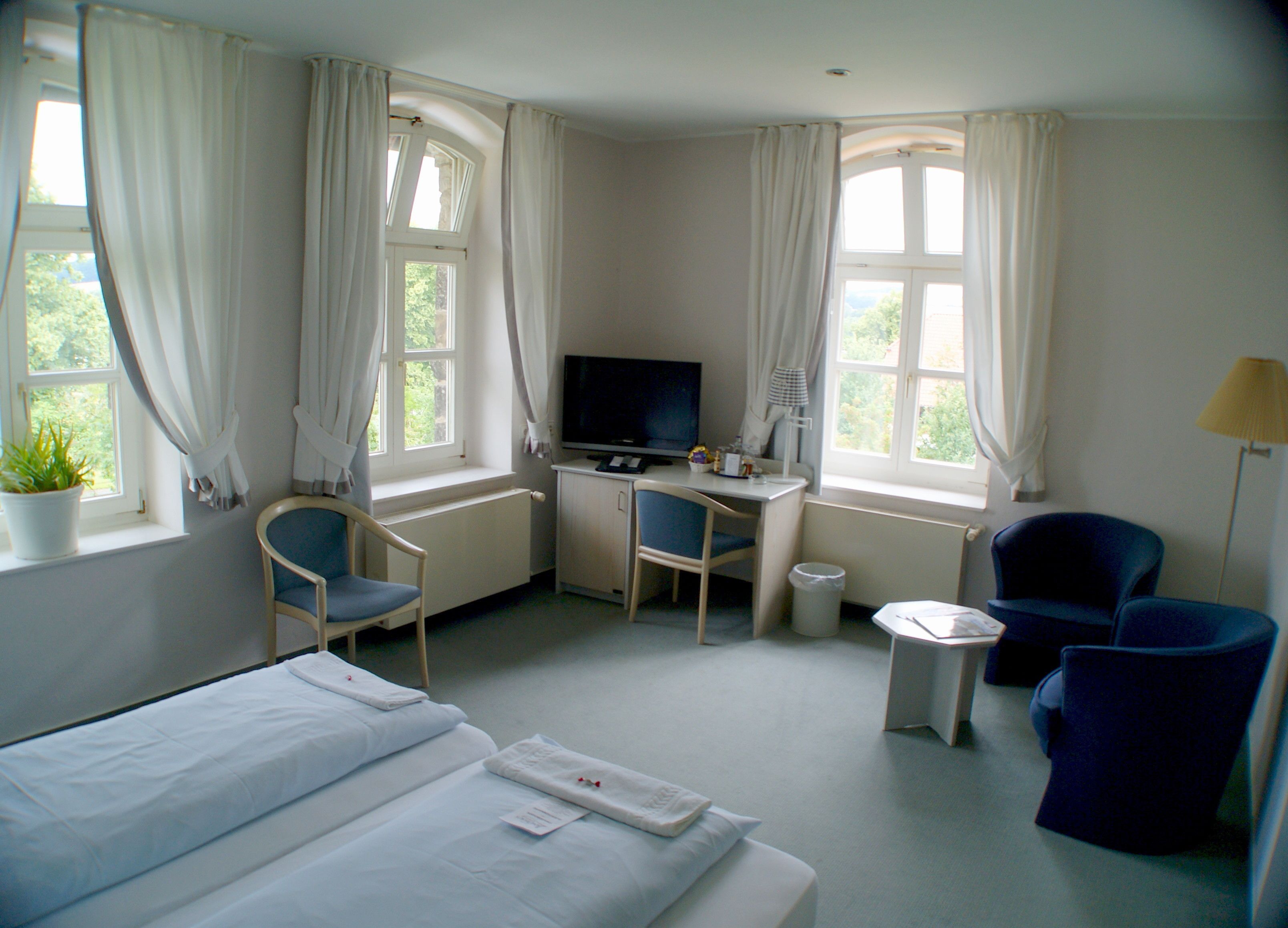 Ein Hotelzimmer mit Bett, Schreibtisch, zwei Stühlen, Fernseher und großen Fenstern mit Vorhängen.