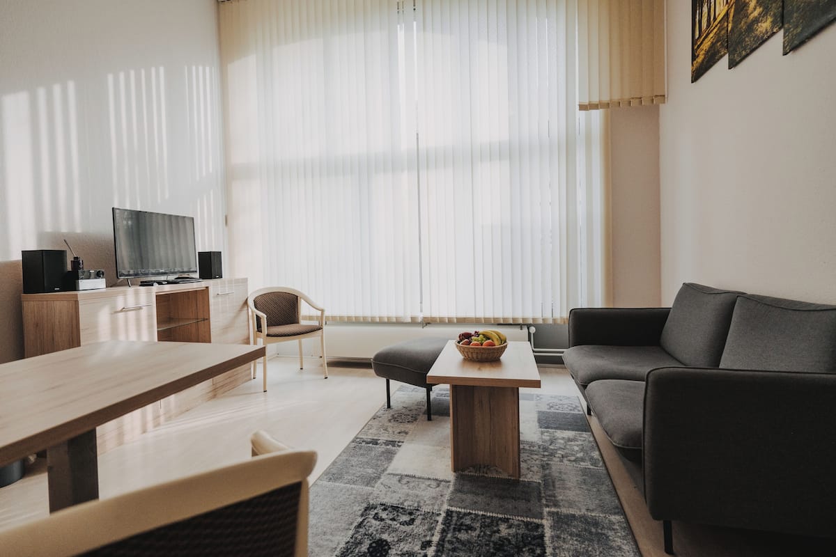 A modern living room with a wooden dining table, a sofa, a coffee table with fruit, a TV, and a window with blinds.