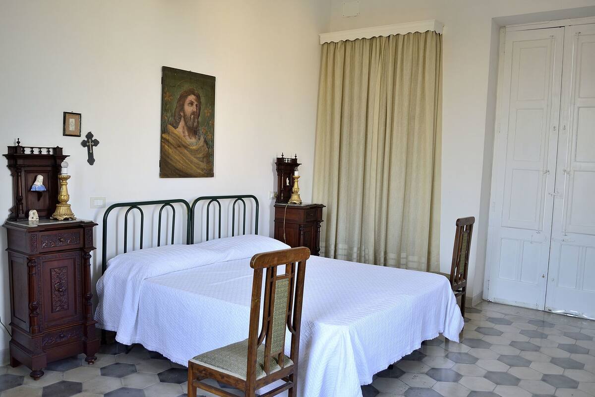 A bedroom with a bed, a chair, a wooden cabinet, a wall-mounted cross, and a painting.