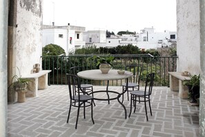 Un balcón con una mesa redonda y sillas, plantas en macetas y vistas a edificios al fondo.
