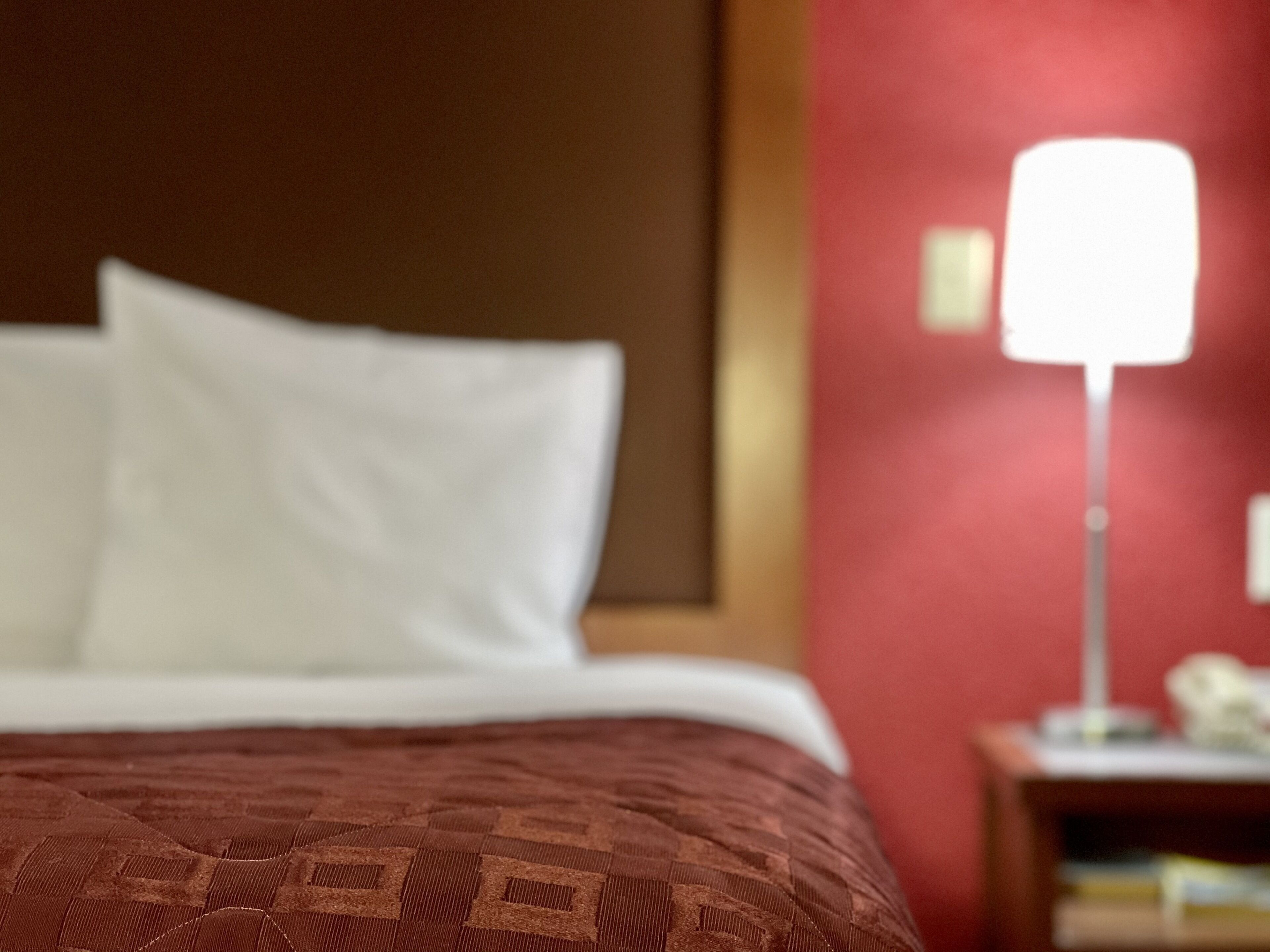 Een bed met een gestreept laken en witte kussens, een rode muur en een nachtkastje met een lamp.