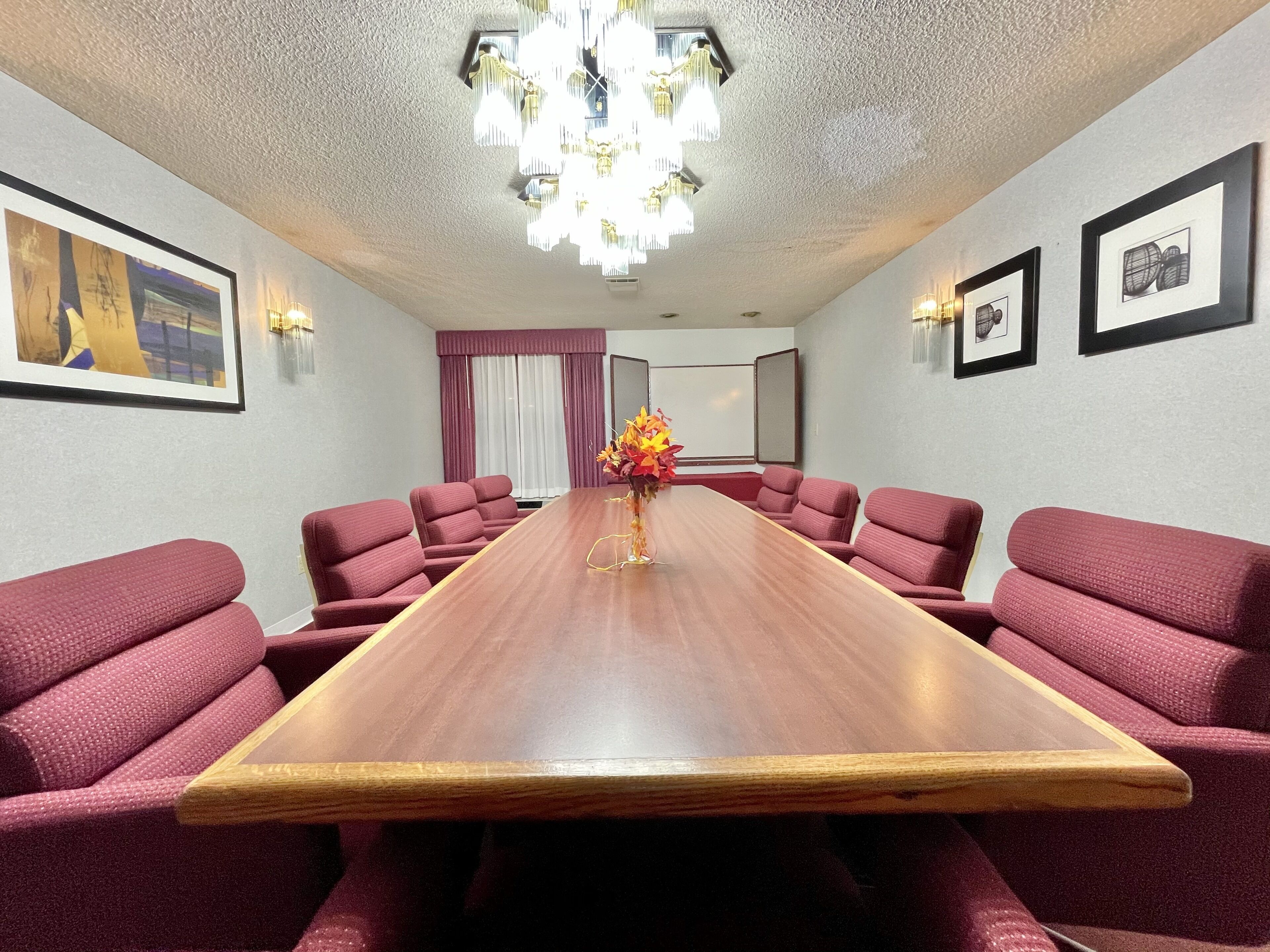 A conference room with a long wooden table, red upholstered chairs, a chandelier, framed artwork on the walls, and a window with curtains.