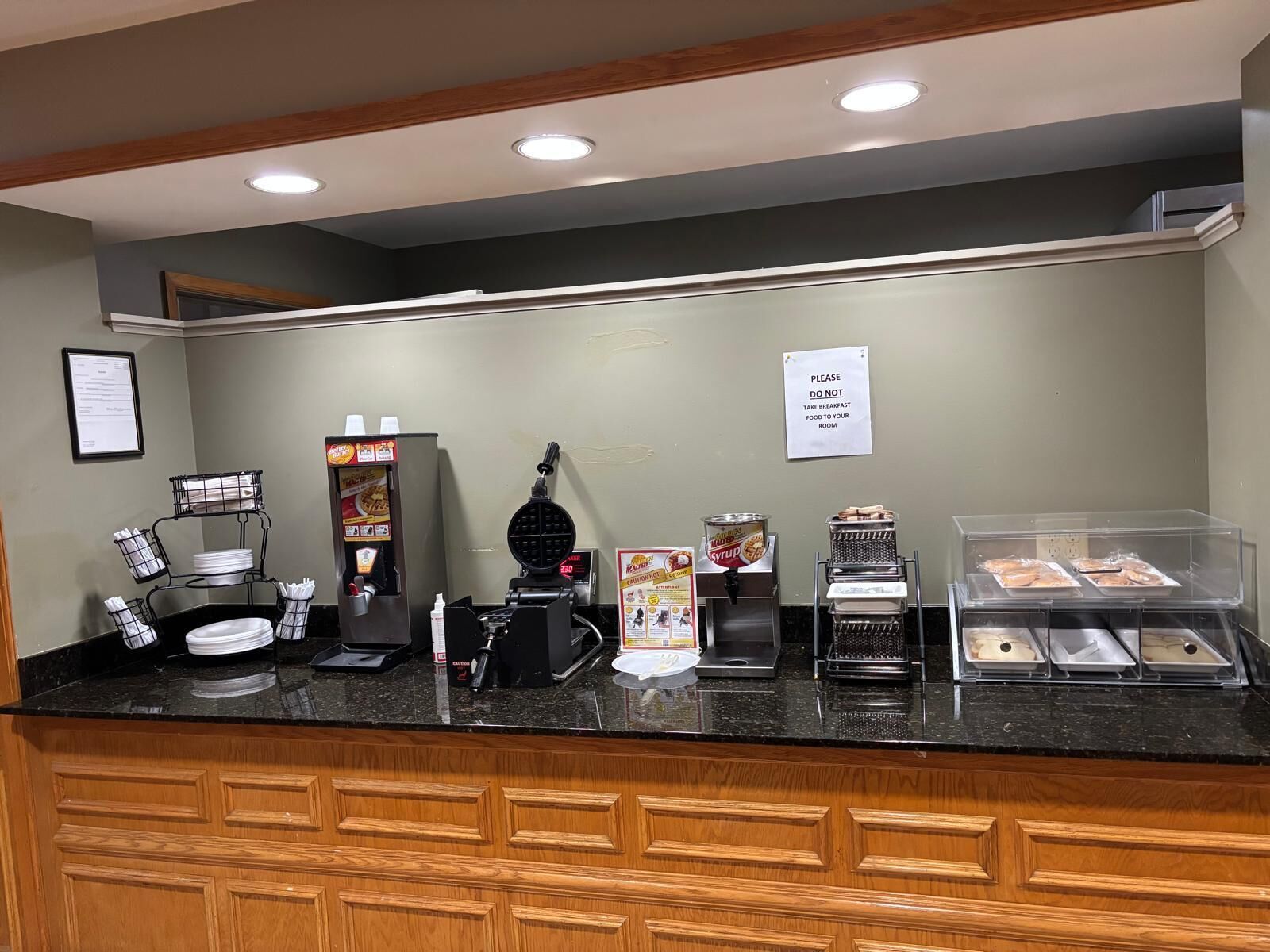 A breakfast counter with a waffle maker, syrup dispenser, and a sign instructing not to take breakfast food to your room.