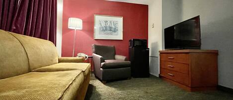 A hotel room with a sofa, armchair, television, and a refrigerator.