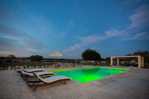 Una piscina con sillones de descanso y una sombrilla, rodeada de muros de piedra y vegetación.