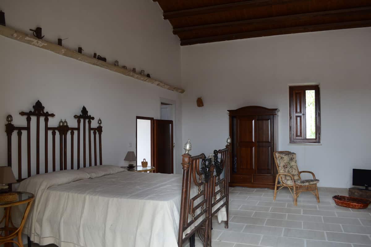Une chambre avec un grand lit, une tête de lit en bois, une chaise en osier, une armoire en bois et une petite table avec une lampe.