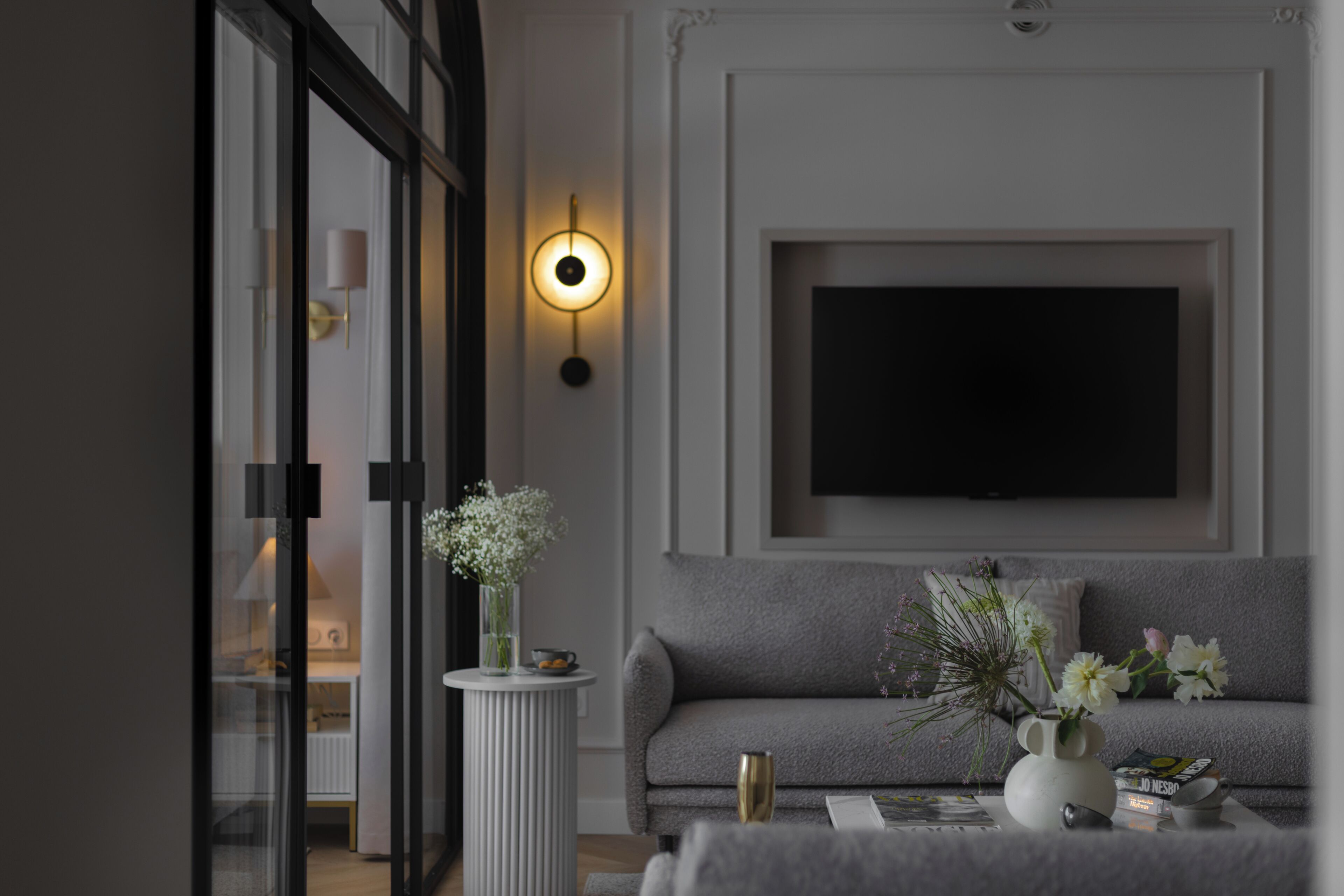 A modern living room with a grey sofa, a flat-screen TV mounted on the wall, a white vase with flowers on a side table, and a glass door leading to another room.