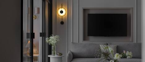 A modern living room with a grey sofa, a flat-screen TV mounted on the wall, a white vase with flowers on a side table, and a glass door leading to another room.