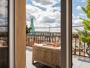 En balkon med en træbænk, en fletkurv med planter, og udsigt til et tårn med et ur.