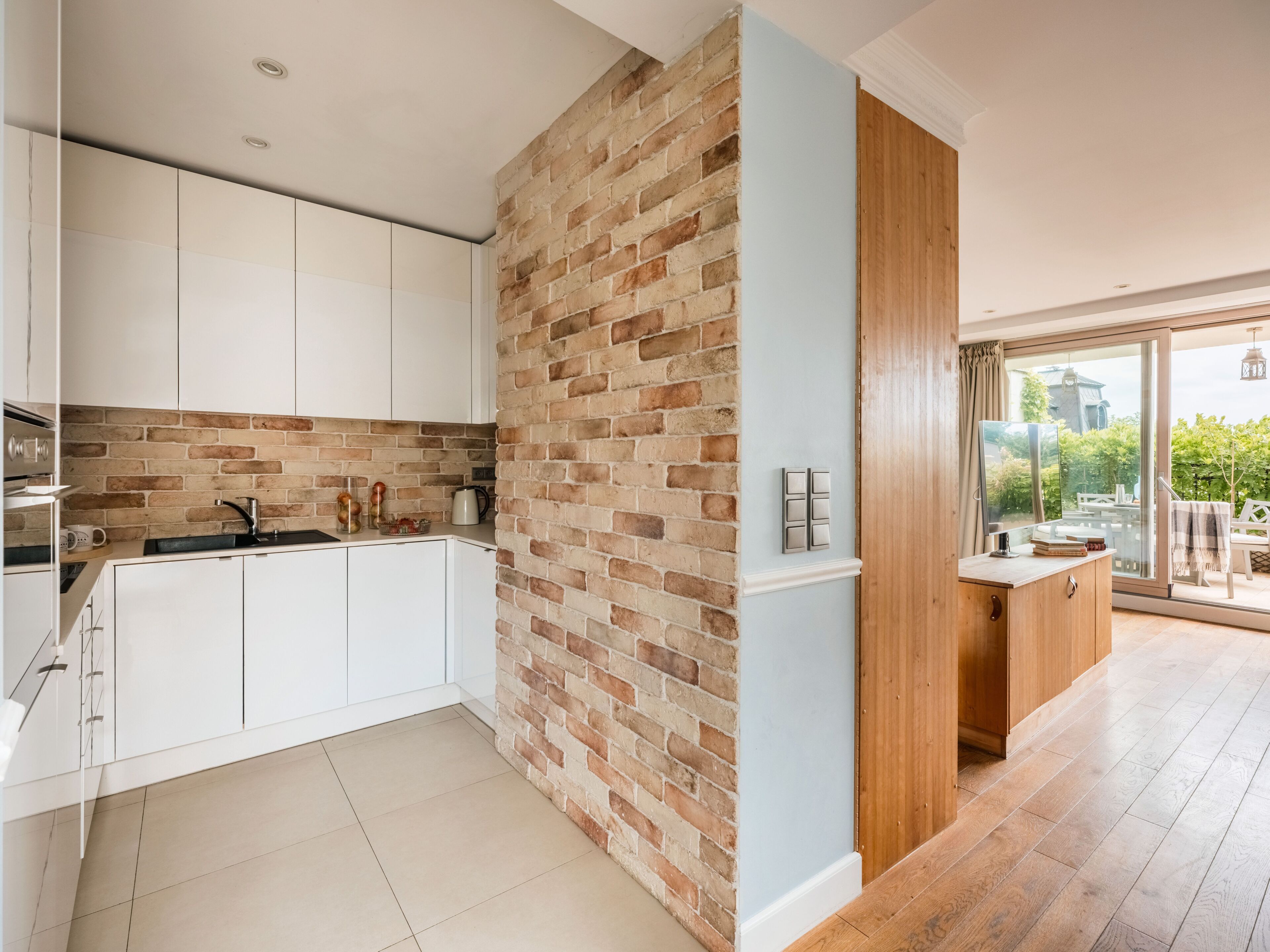 A modern kitchen with white cabinets, a brick accent wall, and a wooden door leading to a balcony.